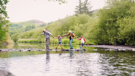 slow motion shot of grandparents helping grandchildren to cross river whilst hiking in uk lake district