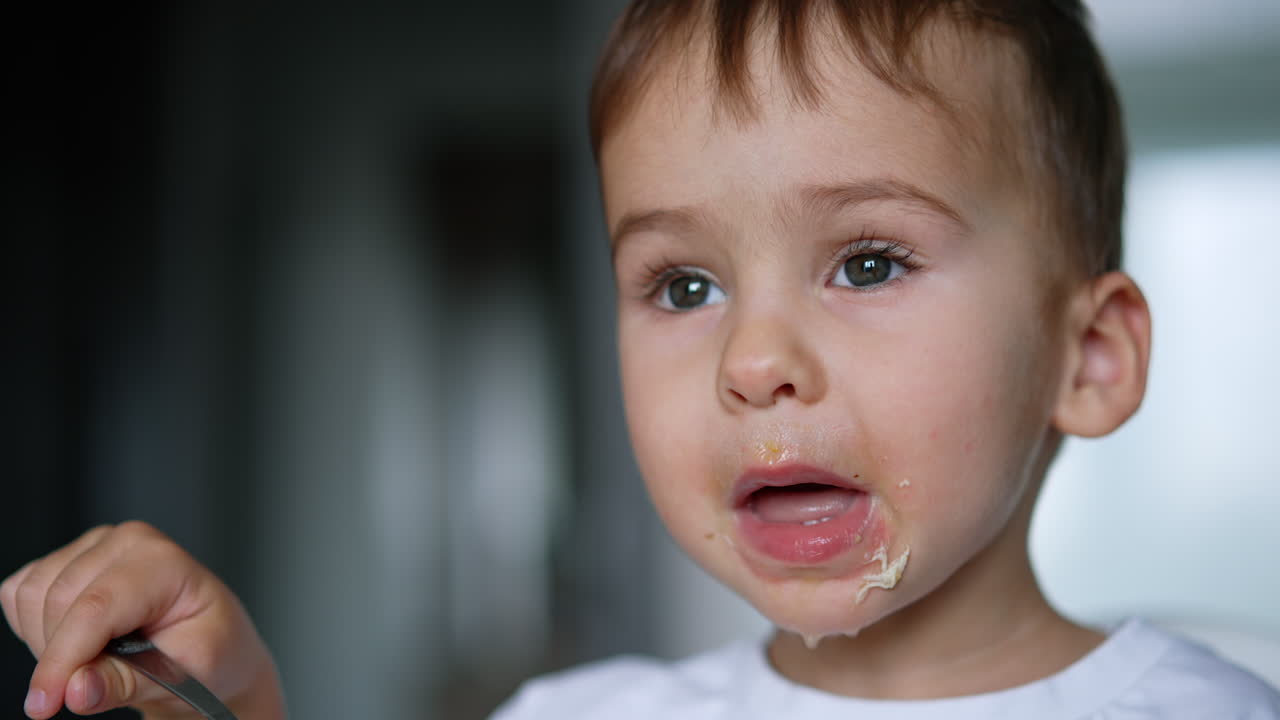 Cute Caucasian toddler enjoying soup. Lovely kid fills the spoon and tastes his food. Close up. Blurred backdrop.