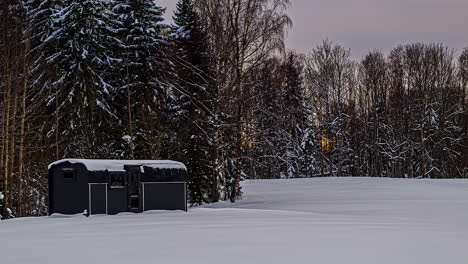 snow-covered-thermowood-cabin-and-leafless-trees-on-winter-scenery,-sunseting-timelapse-view