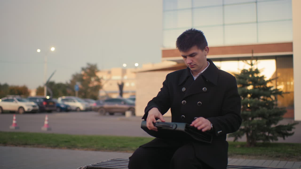 검은색 슈트를 입은 투자자가 벤치에 다가와, 현대적인 건물과 주차된 자동차와 함께 야외 환경에서 브리프케이스를 열고, 새벽에 배경에서 움직이는 자동차, 도시에서 전문적인 비즈니스 순간