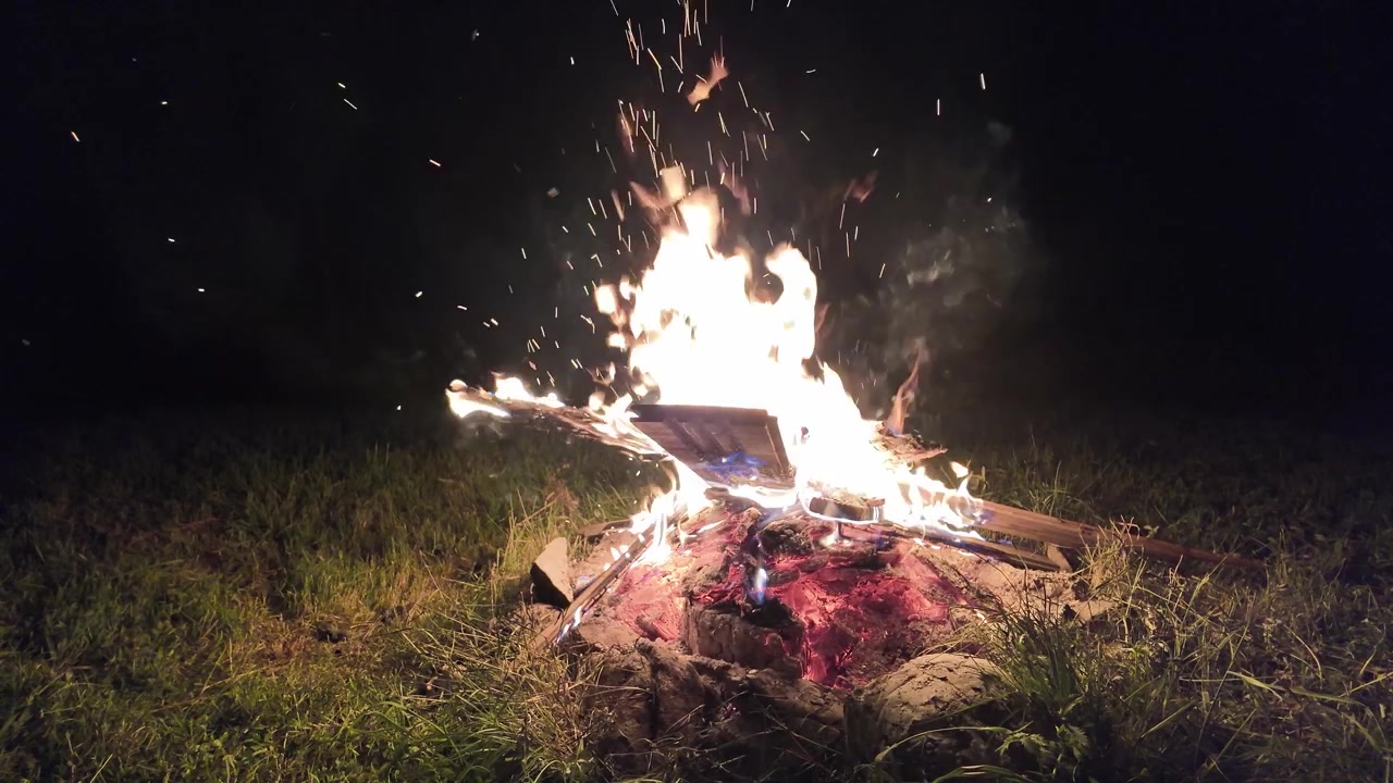 Fogueira brilhante queimando em um prado gramado sob o céu noturno escuro. Brilho quente, brasas voando e ambiente natural ao ar livre, ideal para visuais de viagem, acampamento e aventura.