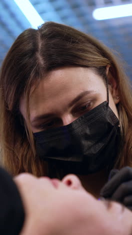 beauty master in black mask uses tool for make up application. female client is having permanent eyebrow make up procedure. low angle view. vertical video