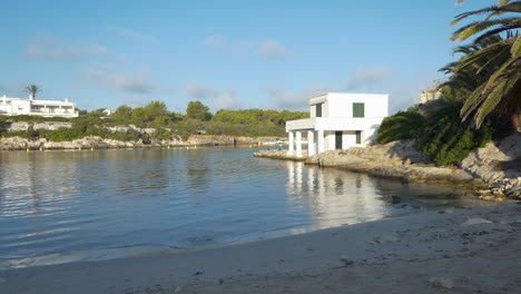 vista lateral al amanecer de la playa vacía en cala santandria en menorca con marcadores de barcos amarillos, mar azul y rocas circundantes.