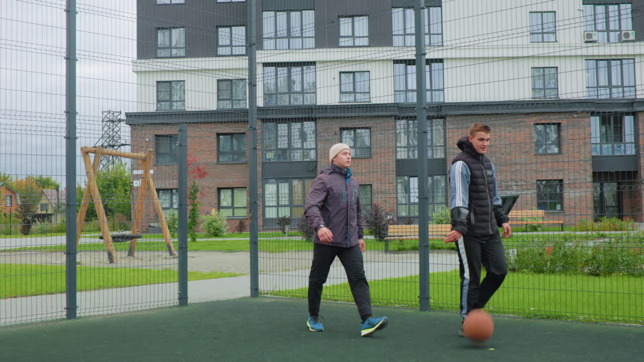 two white men on fenced court holding basketball, focused training scene with coach and trainee dynamic, tactical passing drills and controlled dribble, residential highrise backdrop, cool tones