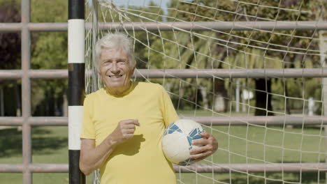 cheerful elderly sportsman standing next to goalpost, holding ball and looking at the camera