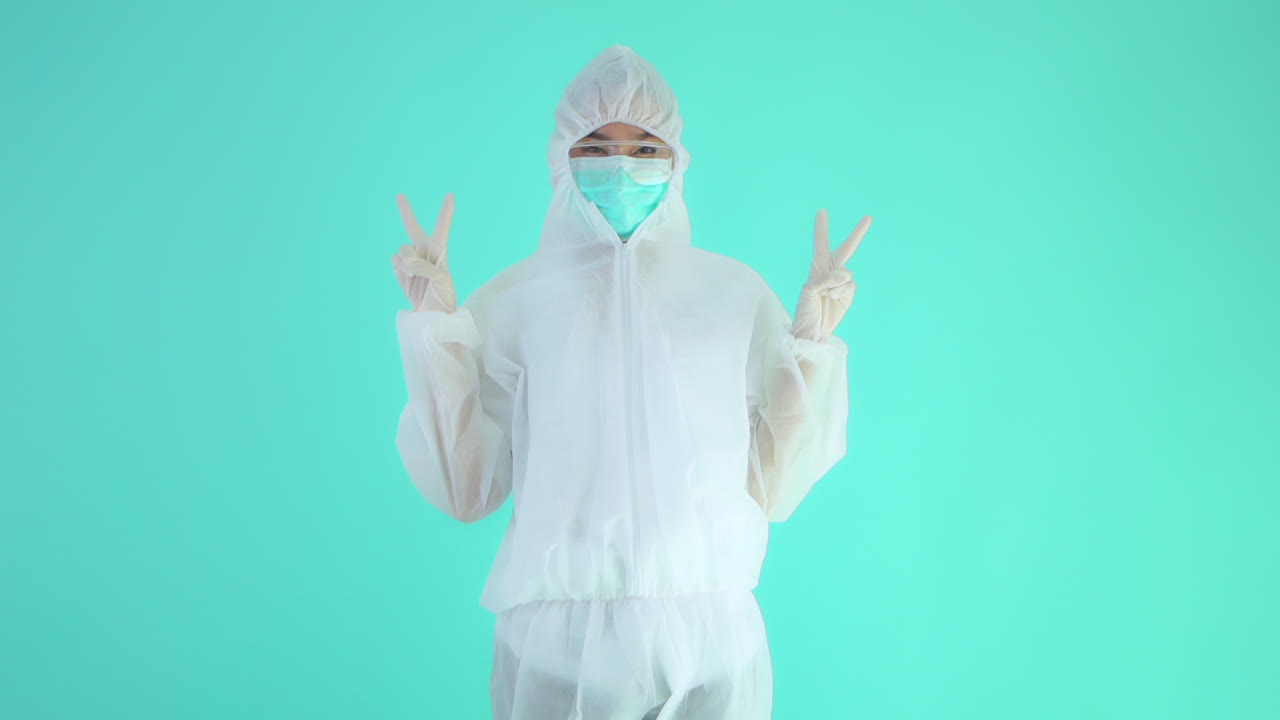 Profissional da saúde usando roupa de proteção contra vírus fazendo sinal de vitória ou paz com os dedos, isolado em um plano de fundo turquesa