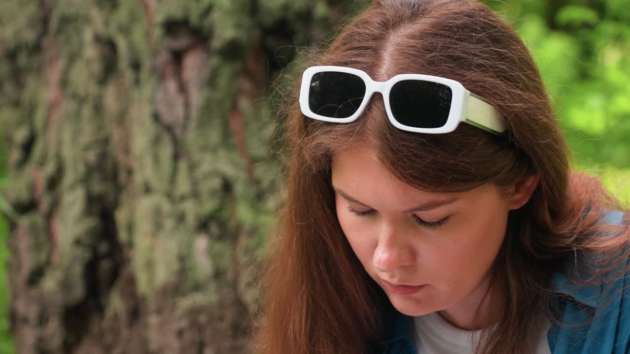 Close up of woman with white sunglasses on head concentrating outdoors, sunlight reflecting on face, wearing white polo and blue shirt, sitting near tree trunk surrounded by lush green nature
