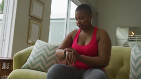 feliz mujer afroamericana de talla grande practicando yoga, usando un reloj inteligente en la sala de estar