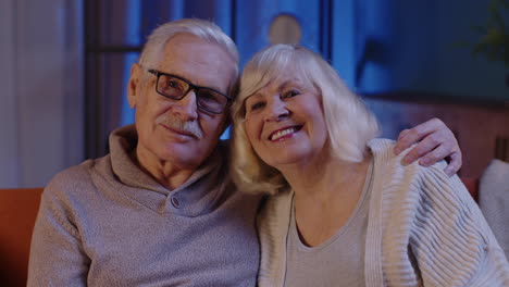 happy old senior elderly family couple hugging, laughing, smiling looking at camera at home sofa