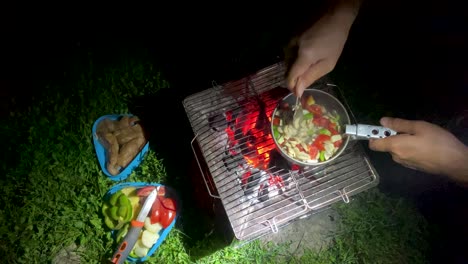 a backpacker man cook sliced vegetable mix with mushroom tomato sauce chicken and veg grill on bonfire wood fire barbecue at night in wilderness survival wild nature in meadow