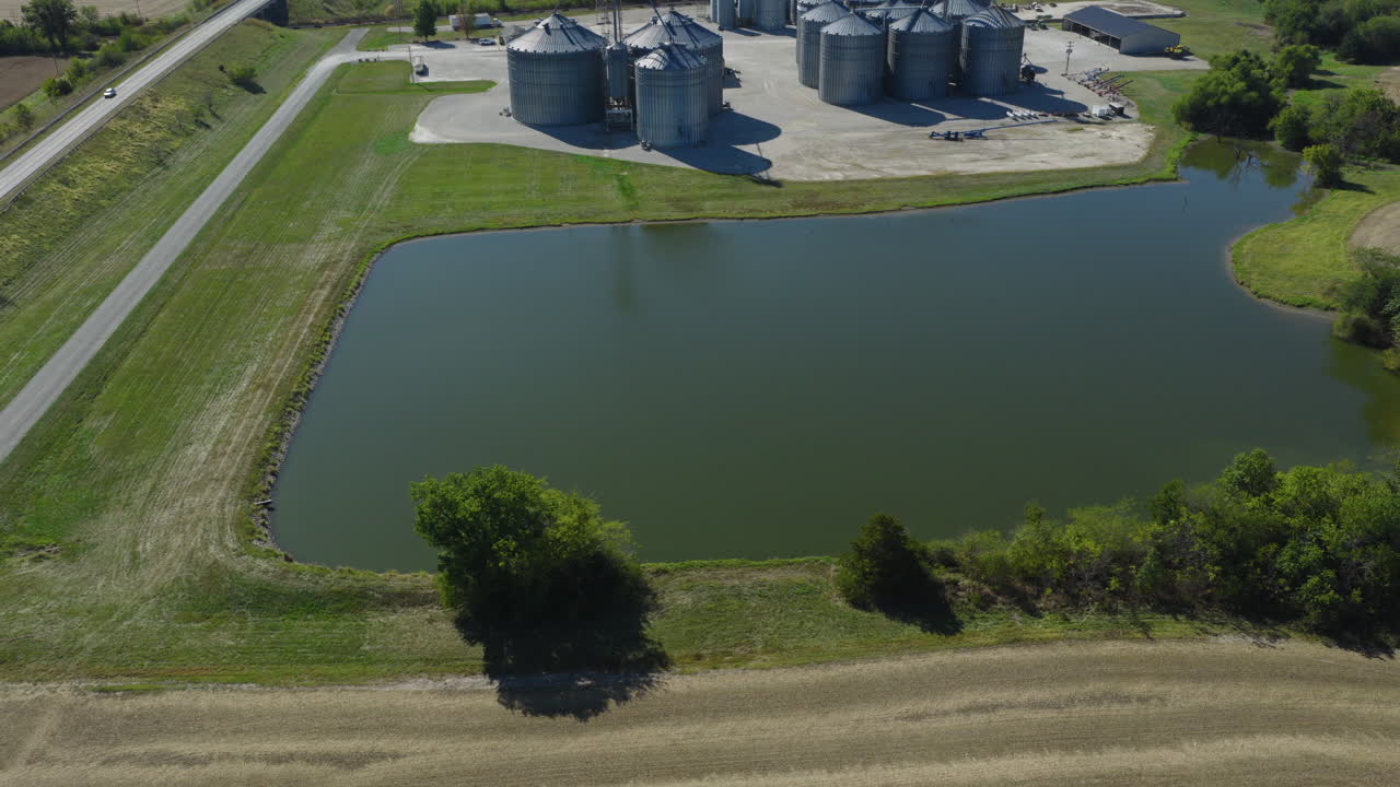 Green retention pond beside farmland, tilt up to grain processing operations and cleared field, aerial pullback