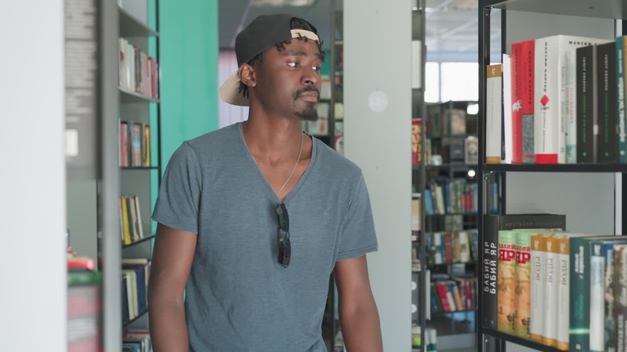 aluno do ensino médio em roupa casual com óculos de sol pendurados na camisa folheia livros na estante da biblioteca, procurando um título em um espaço acadêmico tranquilo com paredes verde-água e fileiras organizadas de literatura