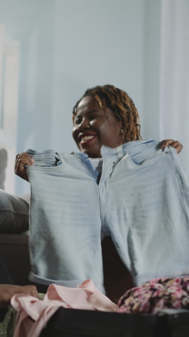 Indian and African American women choose clothes to leave in suitcase for summer trip. Friends enjoy preparing for vacation on seaside sitting on floor
