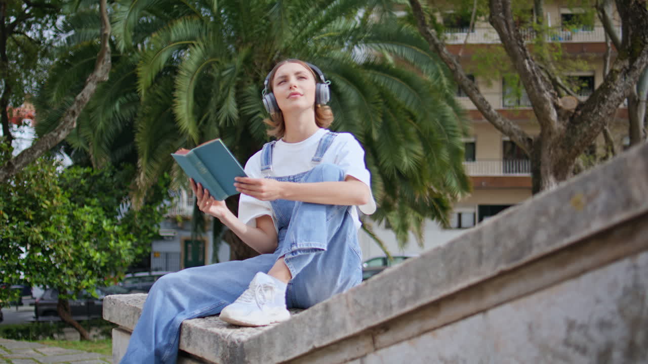 Trendy teenager reading book wearing headphones on stone ledge urban park