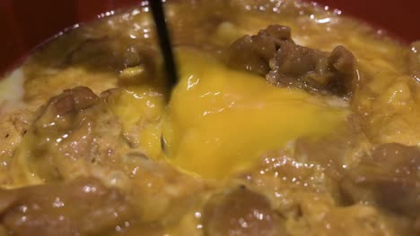 Un-Plato-De-Cocina-Japonesa-Llamado-Gyudon-Se-Mezcla-Con-Palillos-Mientras-La-Yema-De-Huevo-Cruda-O-Un-Huevo-Escalfado-Se-Rompe-Junto-Con-Carne-De-Res,-Cebolla-Y-Arroz-Hervidos