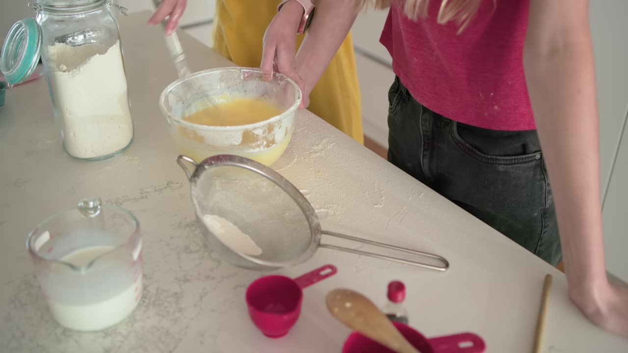 Amigas preparando massa de bolo, despejando leite e batendo a massa em uma tigela na bancada da cozinha.