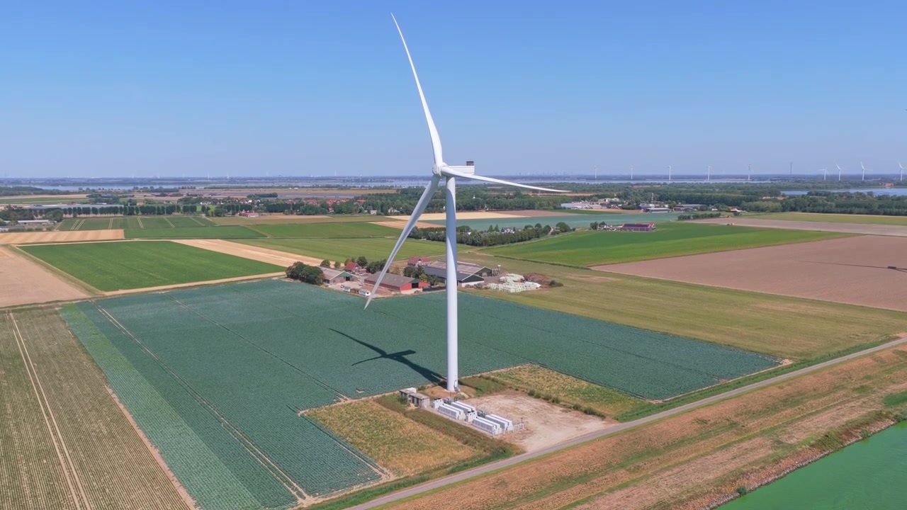 Aerial view of a wind turbine spinning and producing renewable power. A battery energy storage system is visible at the base of the mill