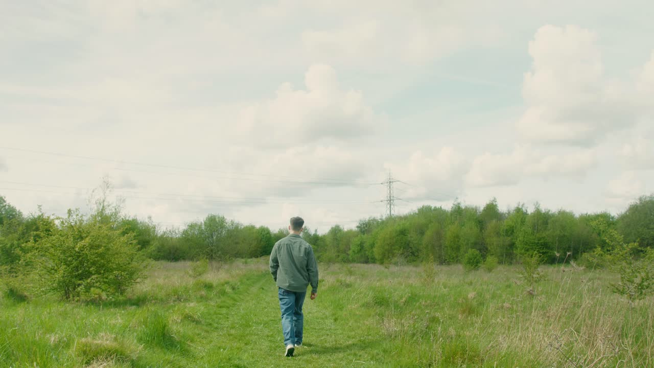 Man walks alone through tall green field under soft cloudy sky, distant landscape fades behind him