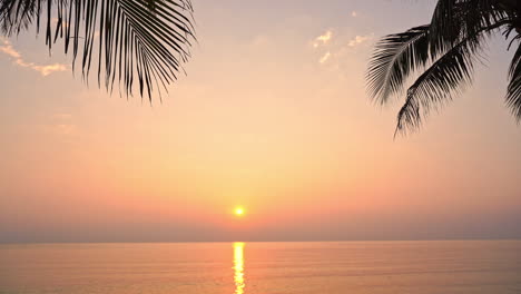 Una-Espectacular-Puesta-De-Sol-Naranja,-Rosa-Y-Amarilla-Sobre-El-Horizonte-Del-Océano-Está-Enmarcada-Por-Hojas-De-Palma