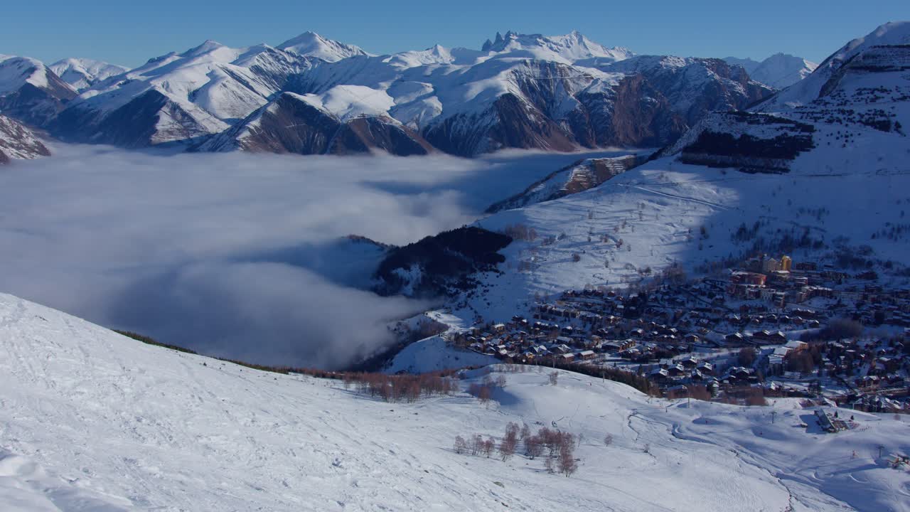 Aerial time lapse of low clouds in the high snowy mountain advancing towards a village on a sunny day