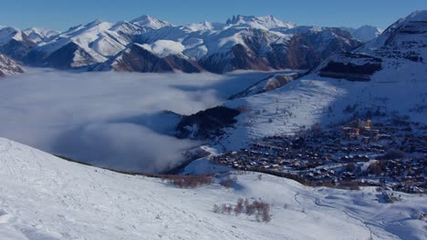 Luftzeitraffer-Von-Niedrigen-Wolken-Im-Hohen-Schneebedeckten-Berg,-Die-An-Einem-Sonnigen-Tag-In-Richtung-Eines-Dorfes-Vorrücken