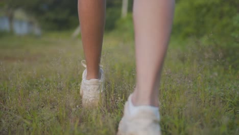 Un-Primer-Plano-De-Pies-Femeninos-En-Zapatillas-Blancas-Caminando-Sobre-Hierba-Verde-Brillante,-Con-Una-Sombra-Proyectada-En-El-Suelo