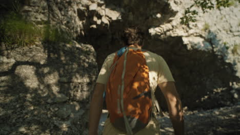 hiker with an orange backpack wearing summer clothes walkig towards the tunnel carved into the rock and entering