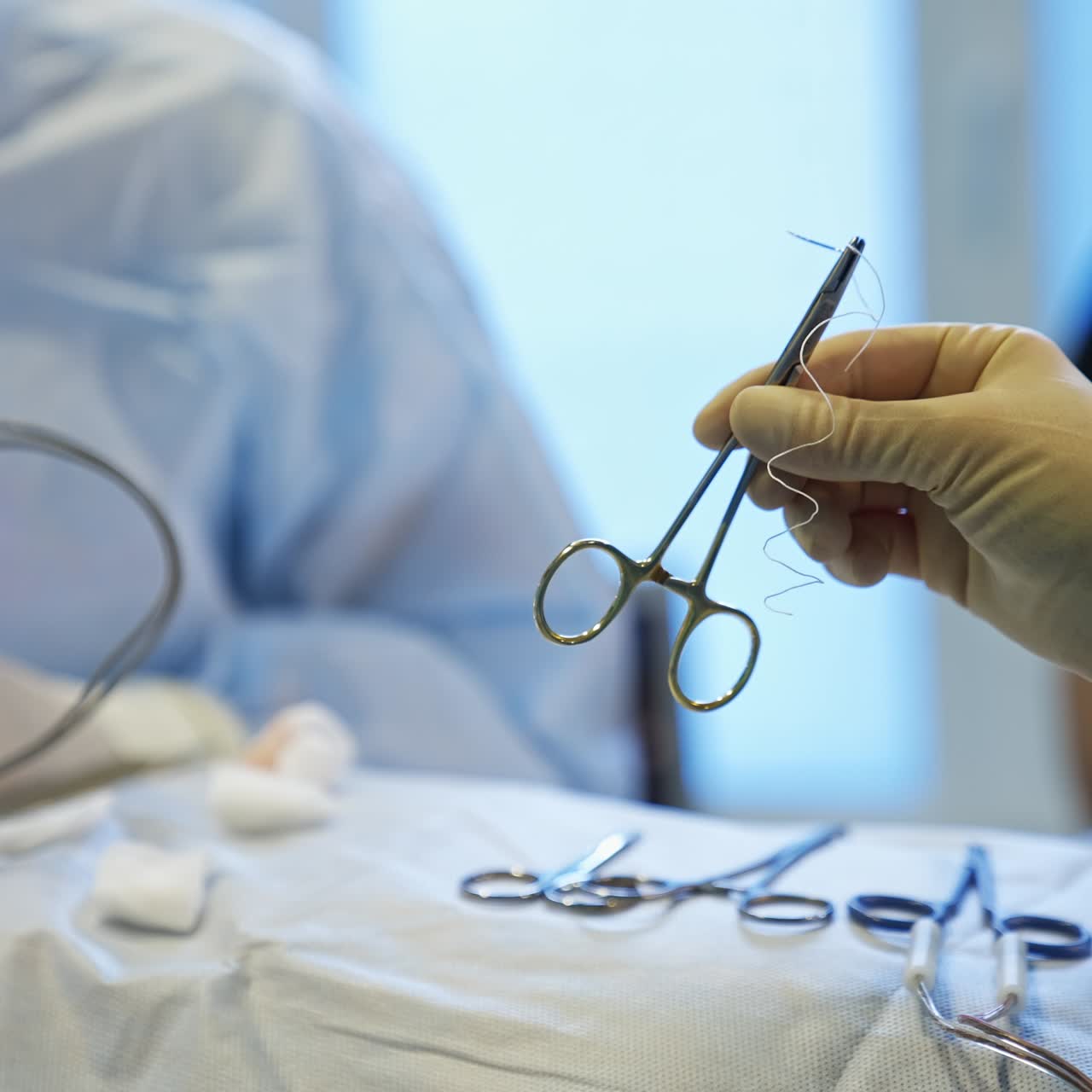 Nurse's hand in latex glove holding the forceps with needle prepared for surgeons' use. Doctors use electric tool at operation. Blurred backdrop