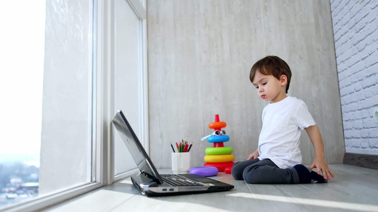 Caucasian kid in white t-shirt sits on the balcony floor. Baby boy looks at laptop and presses keys on the keyboard.