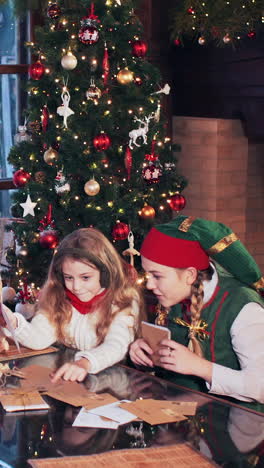 girl and boy are playing at the table with elves in green costumes. santa claus house with christmas tree and fireplace. vertical video