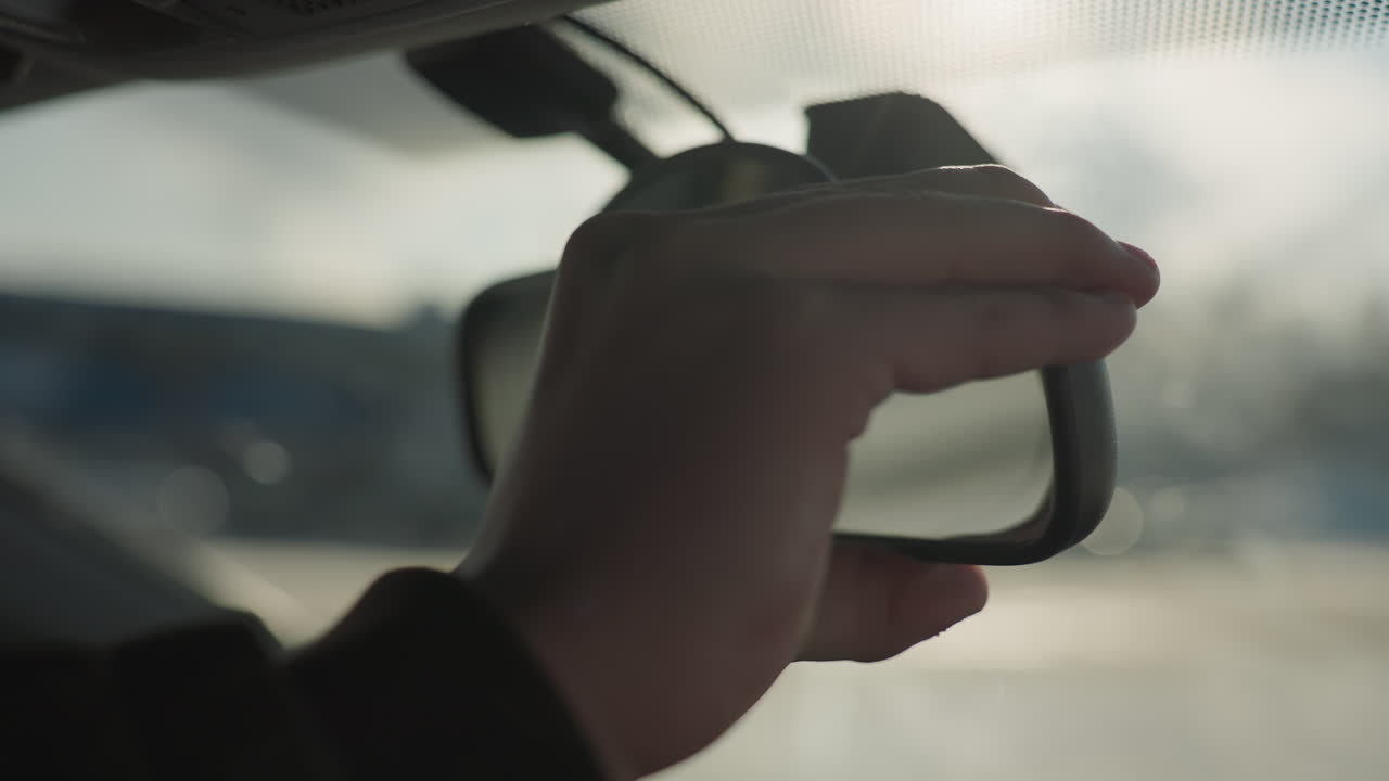 close up hand adjusting rearview mirror inside car against soft blur background capturing warm sunlight lens flare subtle reflection on glass focusing on safety preparation