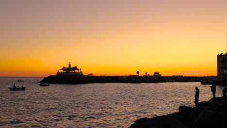 Fishermen-silhouette-on-stony-coast-during-orange-sunset-casting-fishing-line-on-open-sea-in-background-is-city-and-harbor-of-sunset-Grand-Canary-island-valley-4k-slow-motion-capture-at-60fps