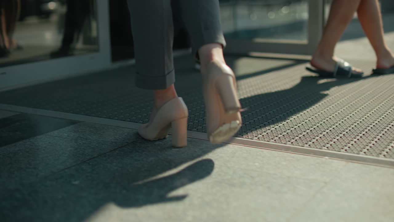 Leg view of woman in high heels walking towards open door on sunny day with people exiting building, shadow cast on ground, focus on movement, footwear, and modern urban professional lifestyle