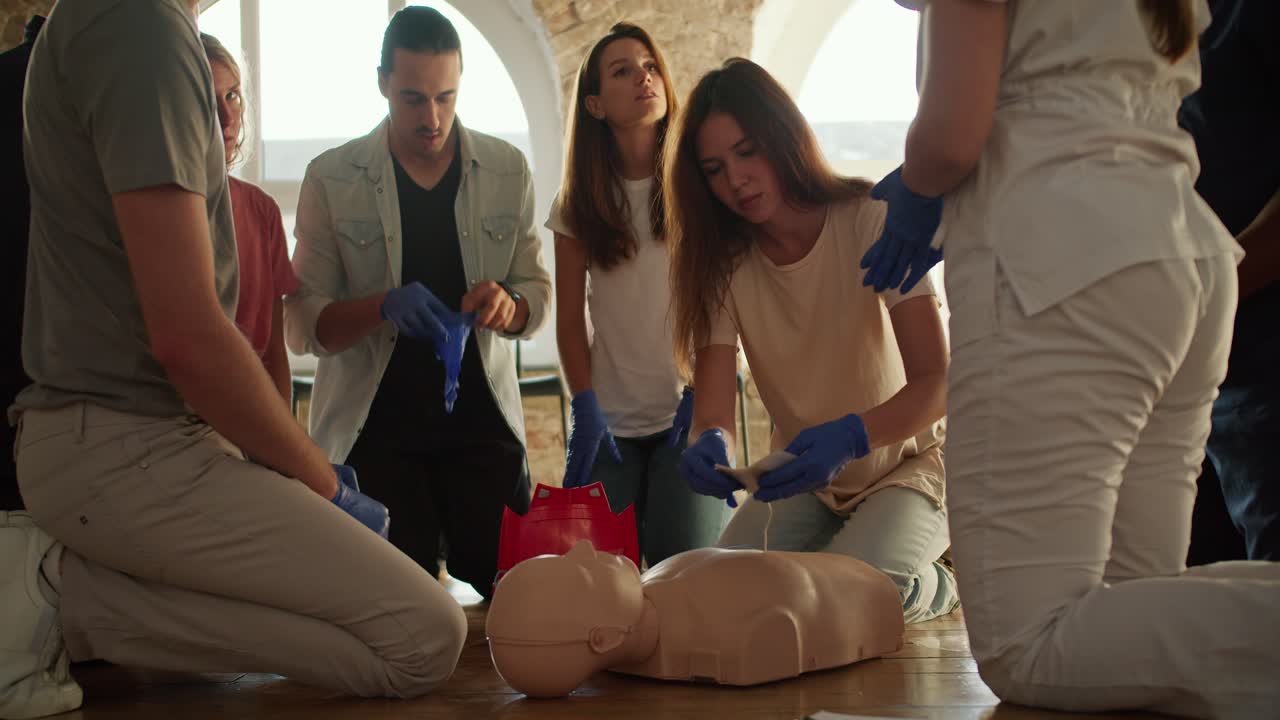 색 티셔츠를 입은 소녀가 전문 의사의 지도하에 마네킨의 몸에 의료 전기 센서를 부착하는 클로즈업 촬영, 그룹의 다른 구성원들이 의사의 설명을 듣는 동안 색 유니폼을 입는 소녀.