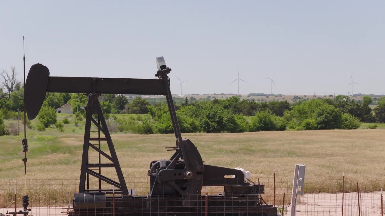 Drone 4K of a wheat field with two oil pump jacks and wind turbines in the background.