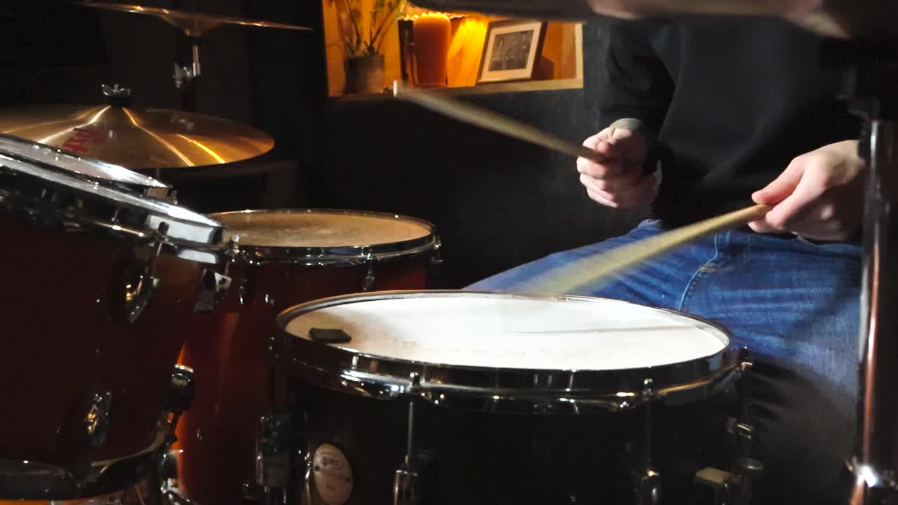 Footage of a man playing acoustic drums or drum set in slow motion in a studio room while hitting the drums and the drumskins with wooden sticks hard. Drums are in a studio room, reflective and glossy