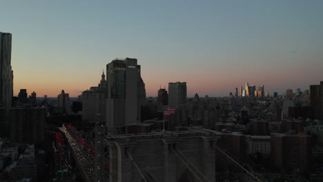 Fly-around-American-flag-raised-on-top-of-bridge-tower.-Busy-multilane-road-in-city-at-dust-and-downtown-skyscrapers-reflecting-setting-sun-in-distance.-Manhattan,-New-York-City,-USA