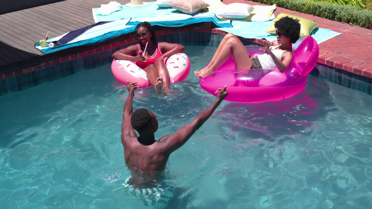 African American friends laughing on donut float as man raising arms to play in backyard pool