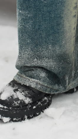 primer plano de piernas en botas negras cubiertas de nieve tocando rítmicamente en el suelo blanco, vaqueros de vaqueros salpicados de helada, capturando un momento invernal de movimiento sutil en medio de un entorno sereno nevado