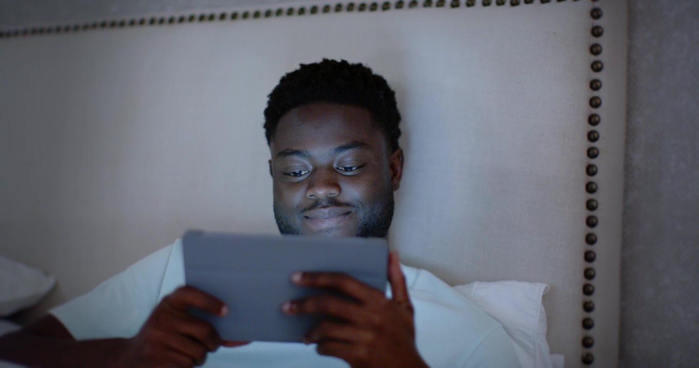 African American adult man reclining on bed holding tablet, blue screen causing scrolling and smile