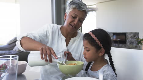 Eine-Gemischtrassige-Großmutter-Gießt-Milch-In-Eine-Schüssel-Für-Eine-Junge-Gemischtrassige-Enkelin-An-Einem-Küchentisch