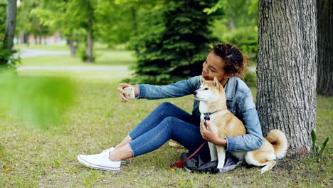 attractive african american girl is taking selfie with cute dog resting in city park cuddling and caressing beautiful animal. modern technology, loving pets and nature concept.