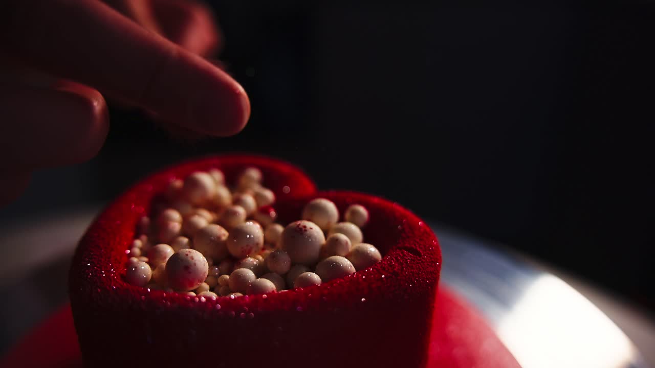 Mano che decora una torta rossa a forma di cuore con zuccherini bianchi