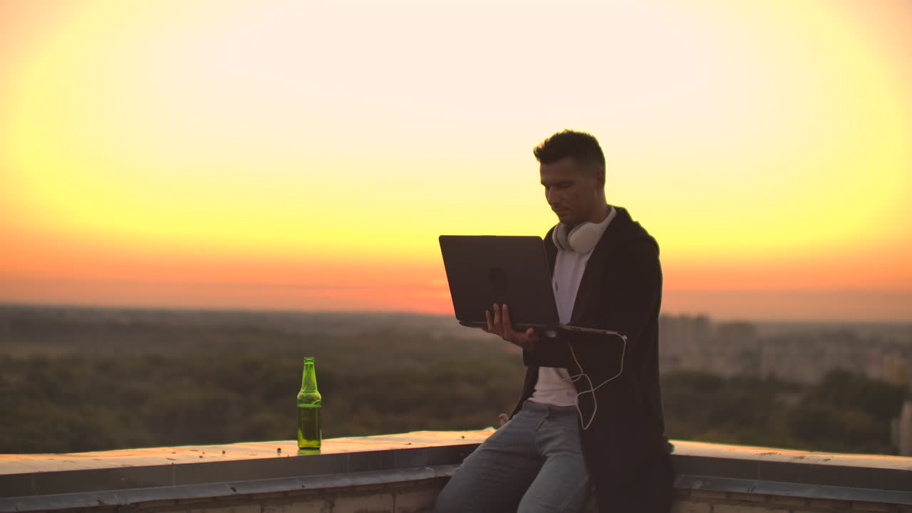 A man in headphones on the roof relaxes working remotely enjoying life despite a handsome kind of sipping beer and types on the keyboard. Trade on the stock exchange using a laptop and enjoying the beautiful view