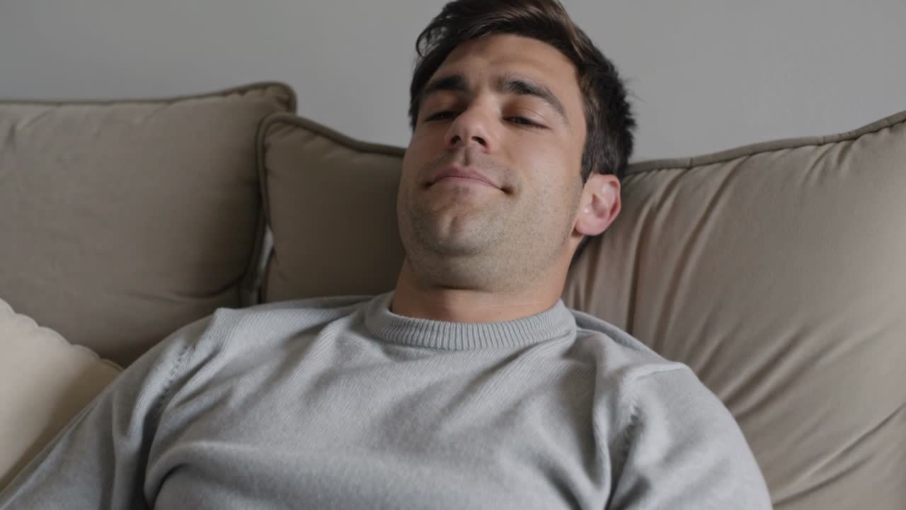 Uomo che si sposta, solleva il mento, apre gli occhi e sorride in cinque fotogrammi su un divano beige con un maglione grigio chiaro