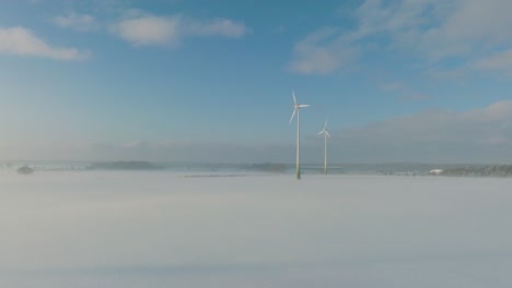 aerial establishing view of wind turbines generating renewable energy in the wind farm, snow filled countryside landscape with fog, sunny winter day, wide ascending drone shot moving forward