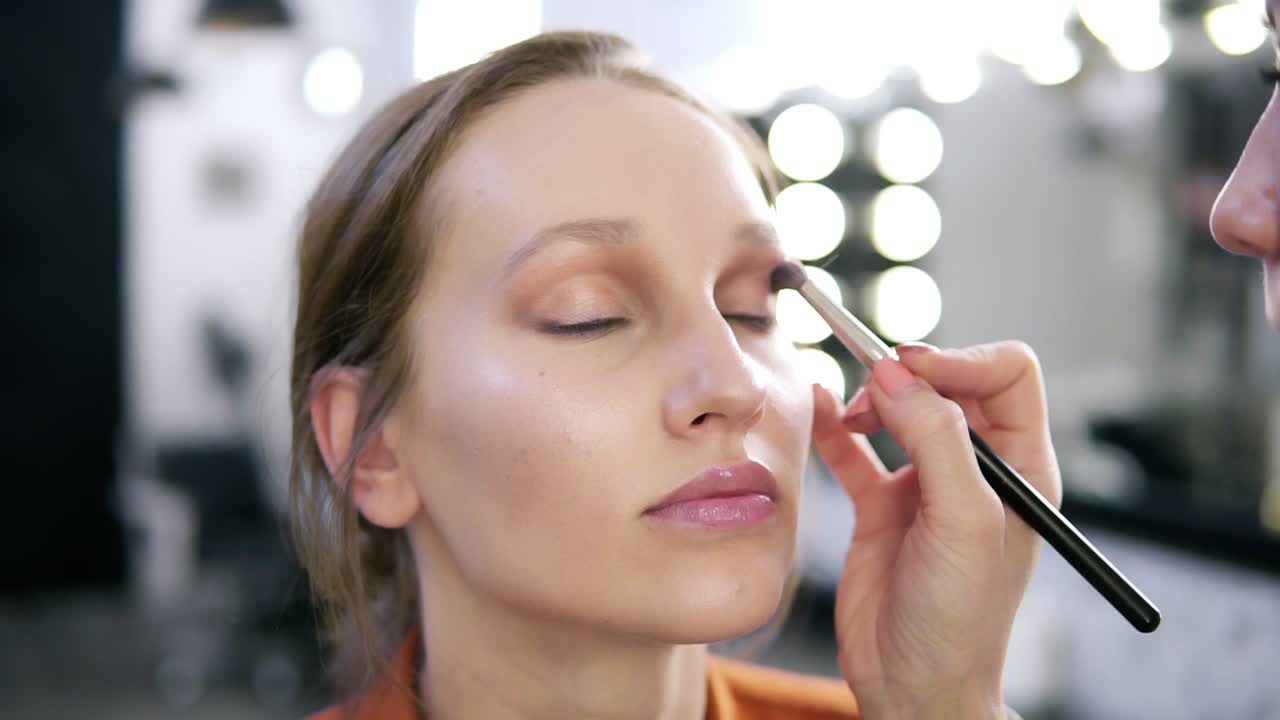 Close up of a model's face during the make up process. Make up master applying light brown eyeshadows. Blurred view of white