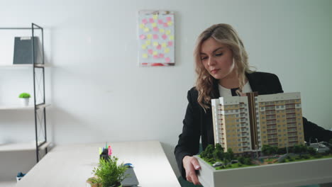 real estate agent lifts architectural building model from desk in brightly lit office, featuring organized shelves with plants and files, green couch, creative sticky note board