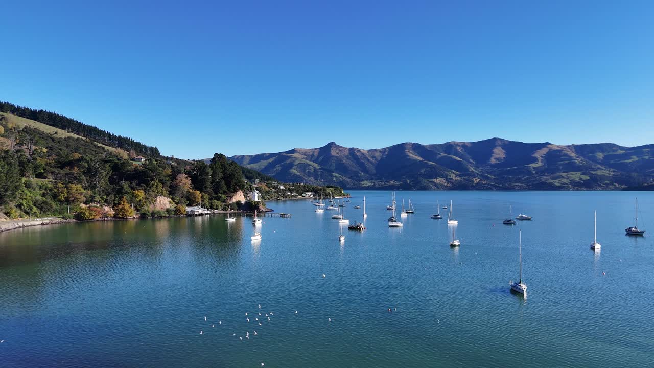Drone footage captures calm waters, sailboats, and distant hills under clear skies in Akaroa, New Zealand