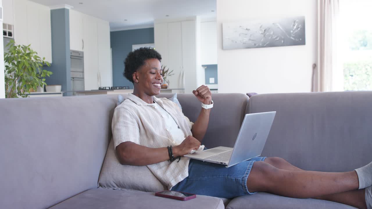 African American man typing on laptop on gray sofa, pausing and pumping fist after success